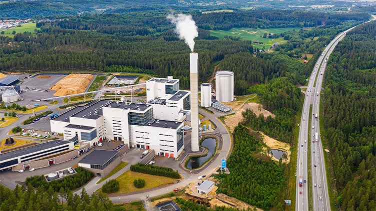 Flygfoto över Jönköping Energis kraftvärmeverk på Torsvik, med vit rök som stiger från skorstenen, omgiven av skog, industriytor och intilliggande motorväg.