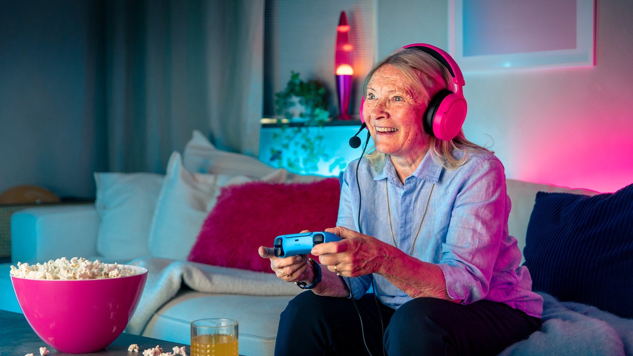 Äldre kvinna med rosa headset spelar tv-spel med handkontroll i ett färgstarkt vardagsrum, med popcorn och dryck på bordet framför sig.