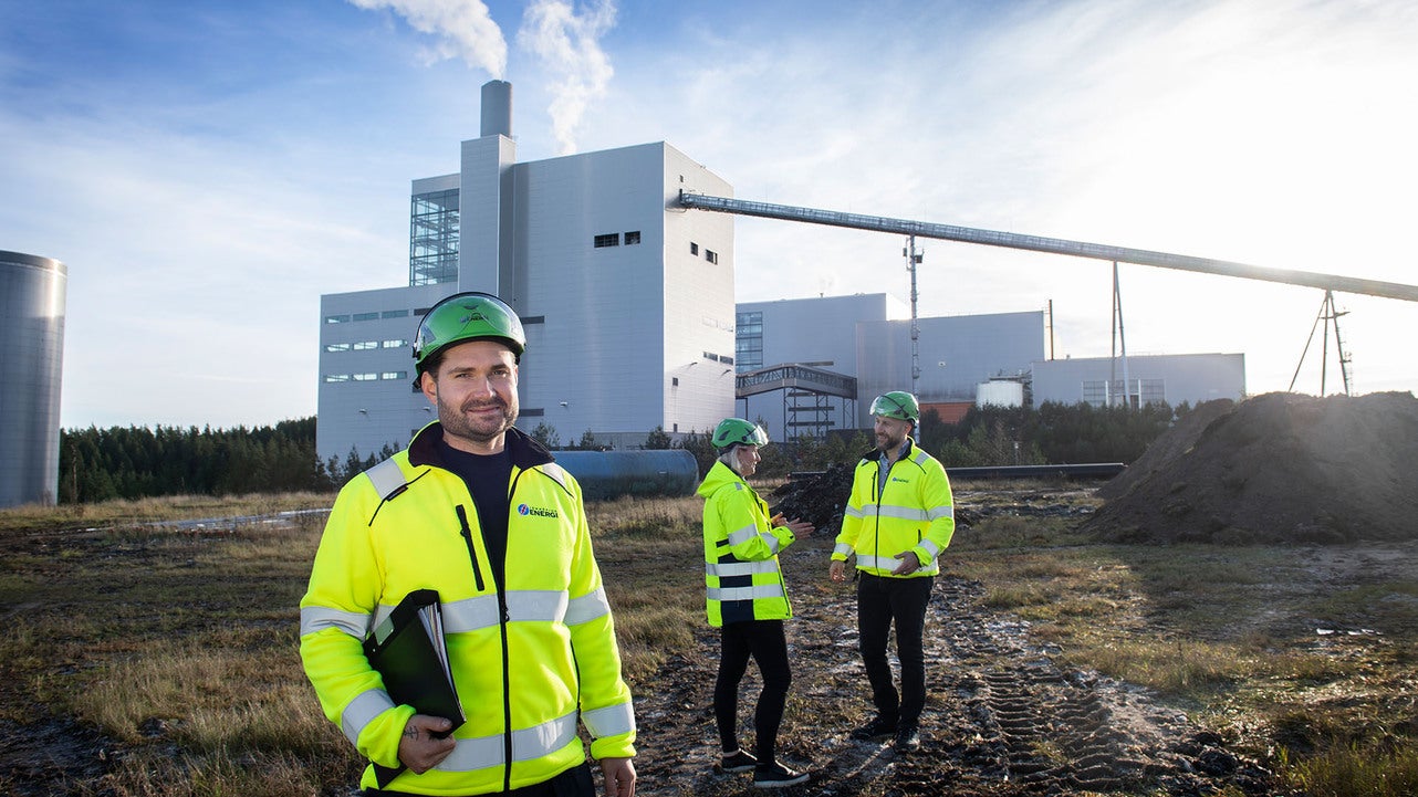 Tre tekniker från Jönköping Energi, klädda i gula skyddsjackor och hjälmar, står utomhus vid kraftvärmeverket på Torsvik. En av dem står i förgrunden och håller en mapp, medan de andra två samtalar i bakgrunden. Rök syns komma från anläggningens skorsten.