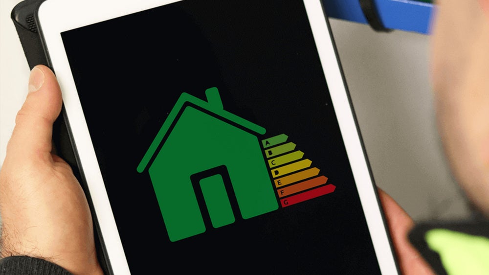 En person håller en surfplatta som visar en illustration av ett hus med en energiklassificering i form av färgade staplar, från grönt till rött, vilket symboliserar energiförbrukning eller energieffektivitet.
