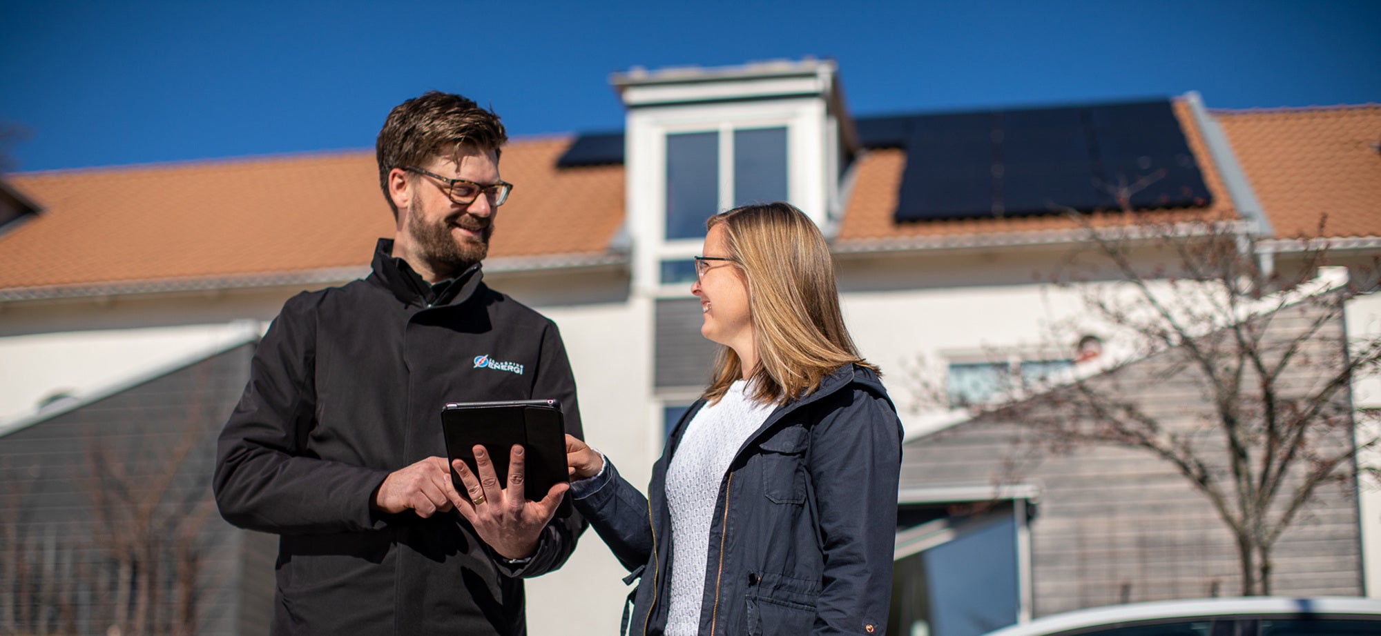 En man i mörk jacka från Jönköping Energi visar en kvinna information på en surfplatta utanför en villa med solpaneler på taket. Solen skiner och båda ler medan de diskuterar.
