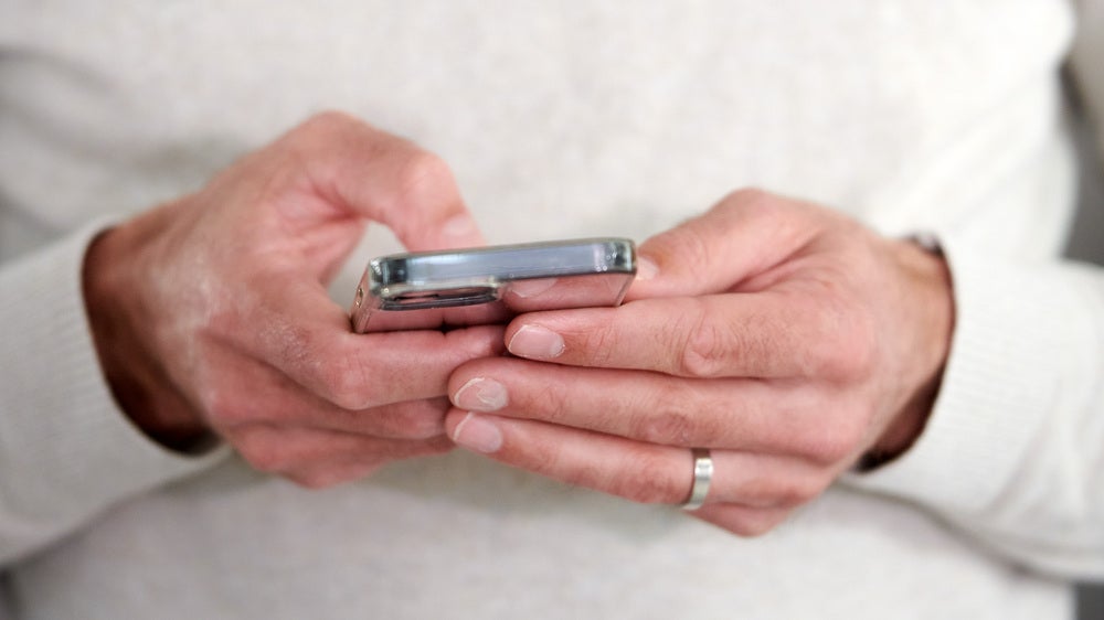 En person i ljus tröja håller en smartphone i båda händerna och tittar på skärmen, med fokus på händerna och telefonen.