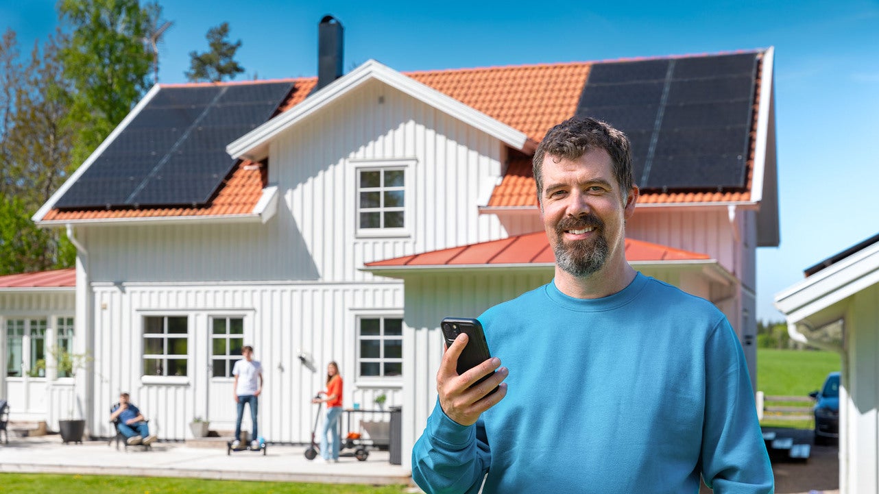 En person står framför ett vitt trähus och håller en smartphone i handen. Huset har solceller på taket och är omgivet av grönska. På utedäcket framför huset sitter flera personer.