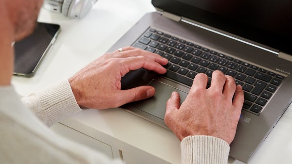 En person sitter vid ett skrivbord och skriver på en öppen laptop. Bredvid datorn ligger en smartphone, och personen är klädd i en ljus tröja.