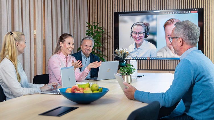 Fyra medarbetare i mötesrum med laptops deltar i ett hybridmöte tillsammans med två kollegor på stor skärm via videolänk