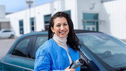 En kvinna i blå jacka står framför en bil och håller i en laddare till elbil.