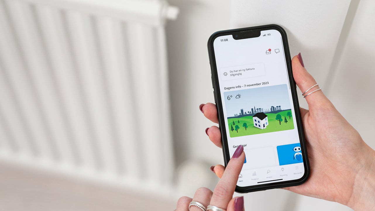 En person använder en mobiltelefon för att hantera sin energiförbrukning via en app. På skärmen syns ikoner som representerar hus och energidata, med ett element suddigt i bakgrunden.
