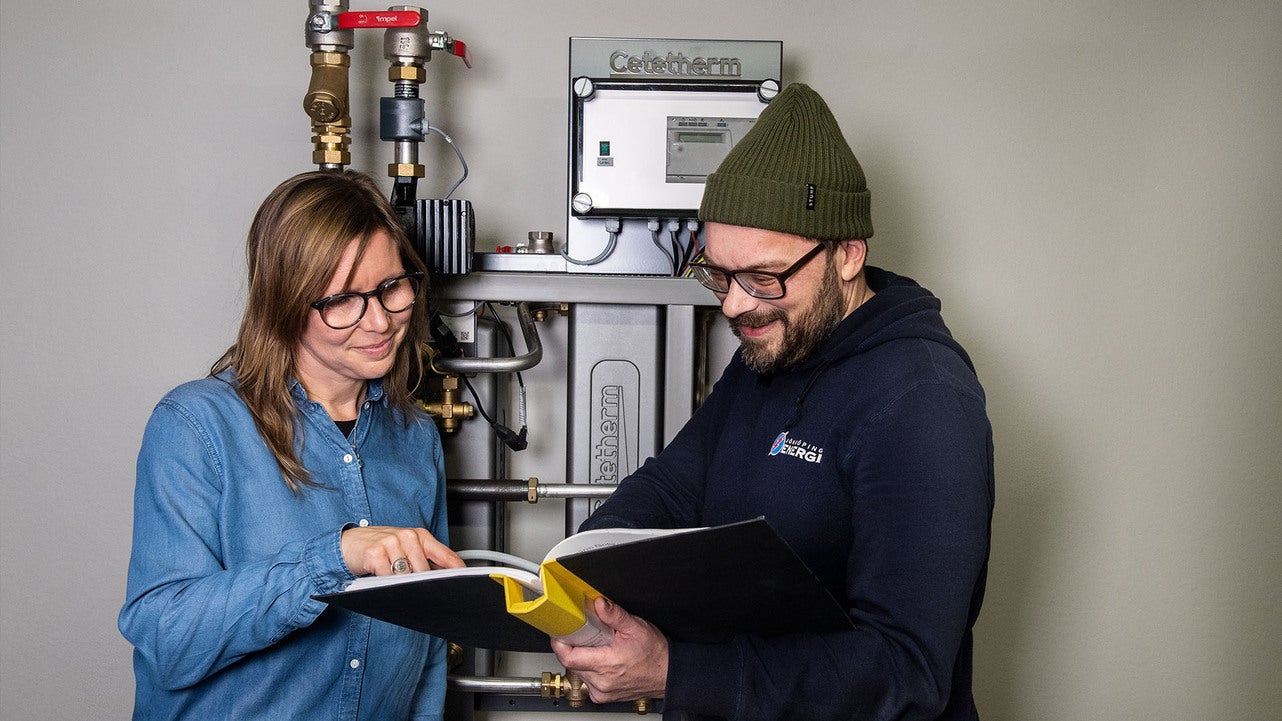 En man och en kvinna står framför ett fjärrvärmesystem och bläddrar i en manual eller teknisk handbok. Fjärrvärmesystemets rör och en kontrollpanel syns tydligt i bakgrunden.