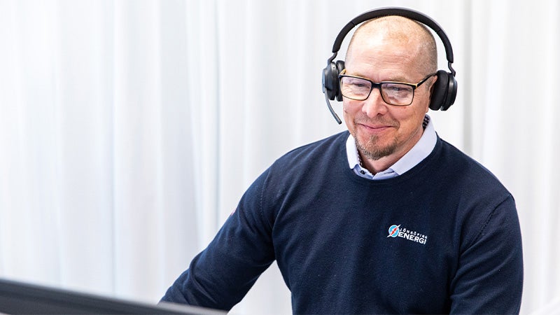 En manlig kundtjänstmedarbetare med glasögon och headset sitter vid en dator, klädd i en mörkblå tröja med företagslogga.