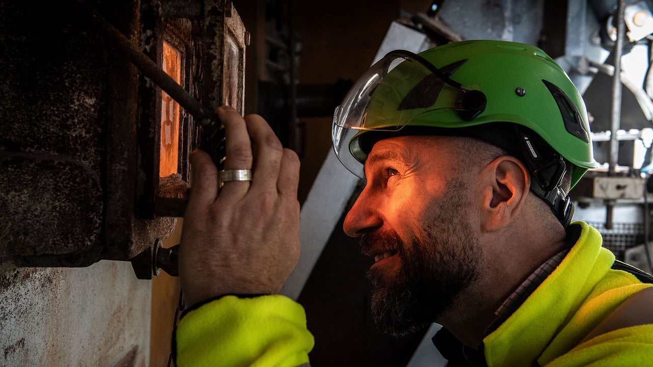 En arbetare med hjälm och skyddsglasögon tittar in i en glödande ugn på en värmeanläggning. Personen bär en gul skyddsjacka och är omgiven av industriell utrustning.