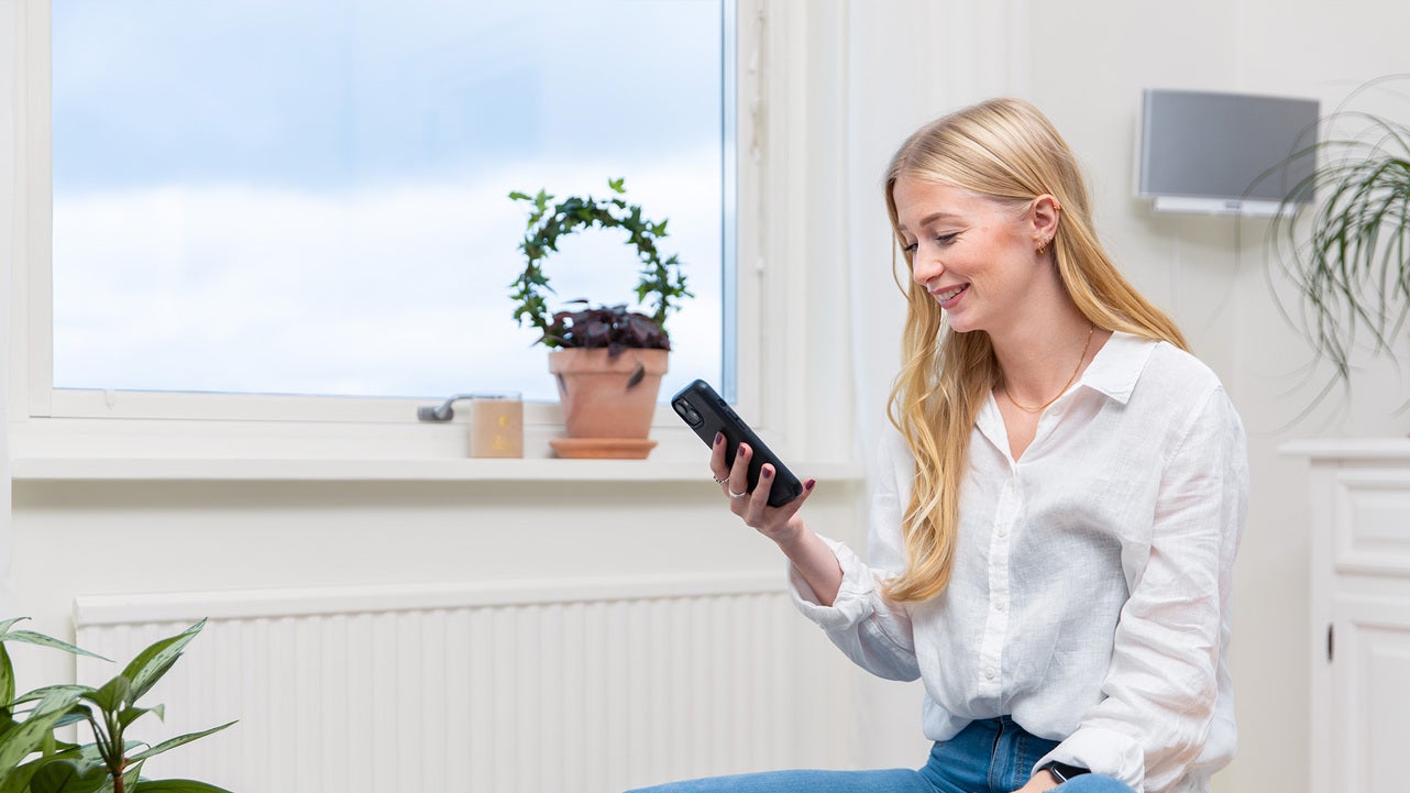 En kvinna med långt blont hår och vit skjorta sitter vid ett fönster och använder en mobiltelefon. I bakgrunden syns ett element och en krukväxt.