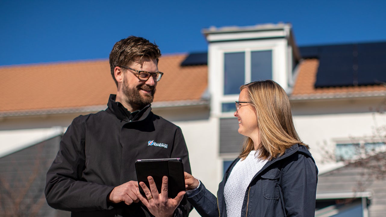 En manlig medarbetare från Jönköping Energi står med en surfplatta och diskuterar med en kvinna framför ett hus med solpaneler på taket. Båda ler medan de tittar på skärmen, och en klarblå himmel syns i bakgrunden.