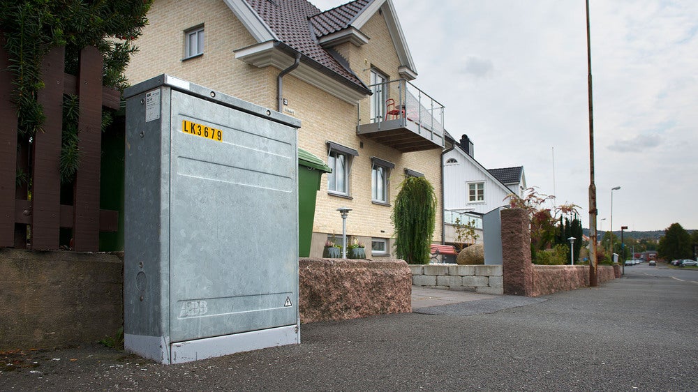 Ett elskåp i metall står längs en trottoar i ett bostadsområde, framför en rad moderna hus. Skåpet är märkt med en gul identifieringsskylt och omges av hushållens soptunnor och trädgårdsdetaljer.