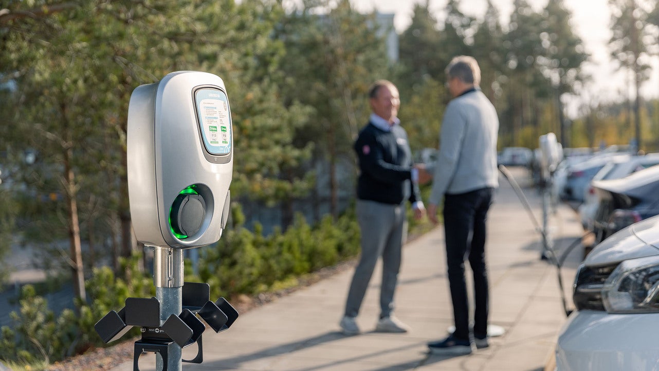 En elbilsladdstation är i fokus på en parkeringsplats, med två personer som står och pratar i bakgrunden. Flera bilar är anslutna till laddstationerna, och scenen utspelar sig utomhus med träd och en solig himmel.