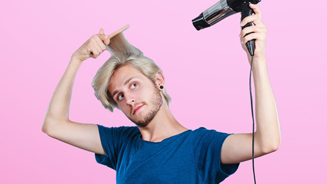 En man med blont hår och blå t-shirt använder en hårfön och kam för att styla sitt hår. Han står framför en rosa bakgrund.