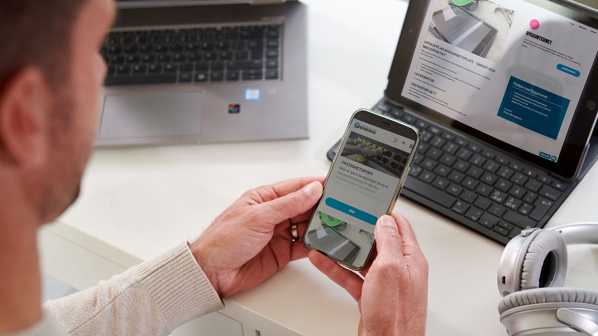 En person sitter vid ett skrivbord och håller en smartphone, medan en surfplatta och en bärbar dator är öppna bredvid. Personen använder flera enheter samtidigt för att läsa och arbeta.
