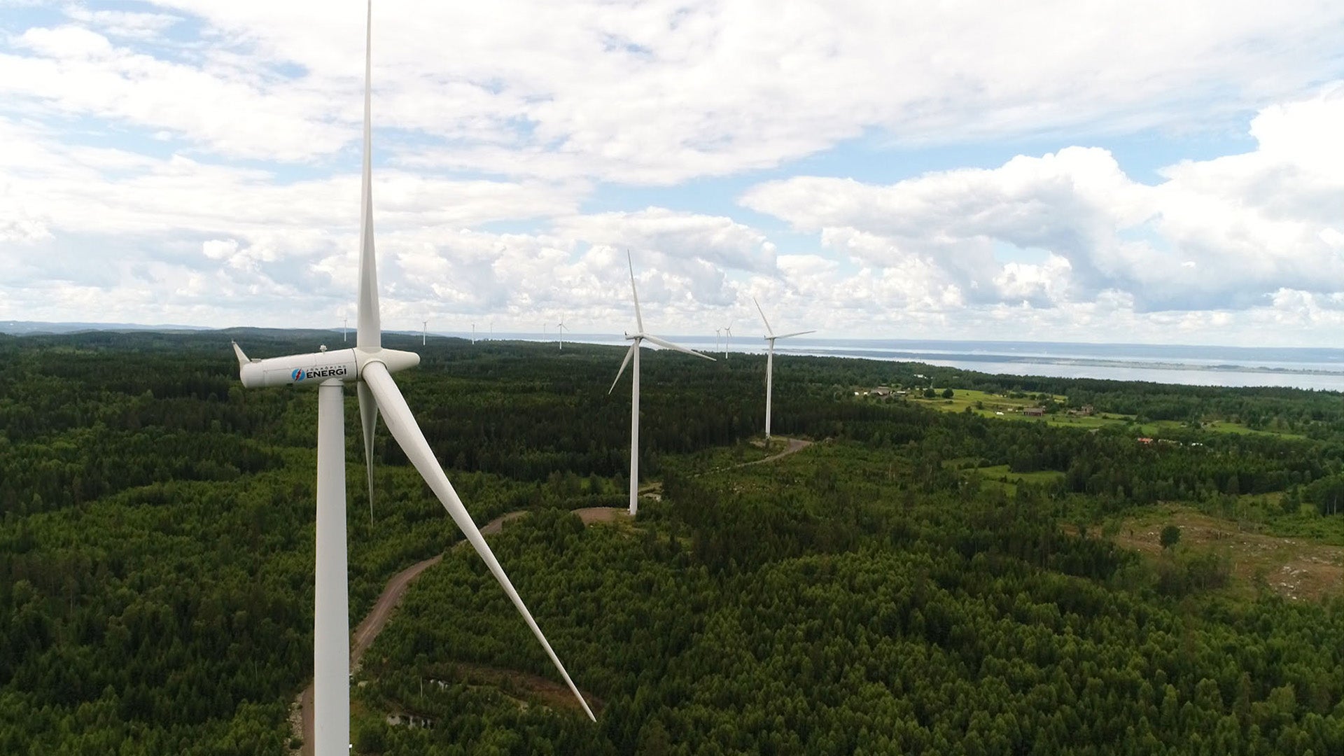 Flera vindkraftverk reser sig över ett grönt skogslandskap med sjön Vättern i bakgrunden under en molnig himmel.