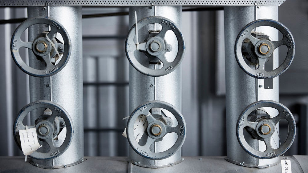 En rad av sex industriella ventiler monterade på stora metallrör i en teknisk anläggning. Ventilerna har handtag som används för att styra flödet i systemet, och rören är märkta för att indikera deras funktion.