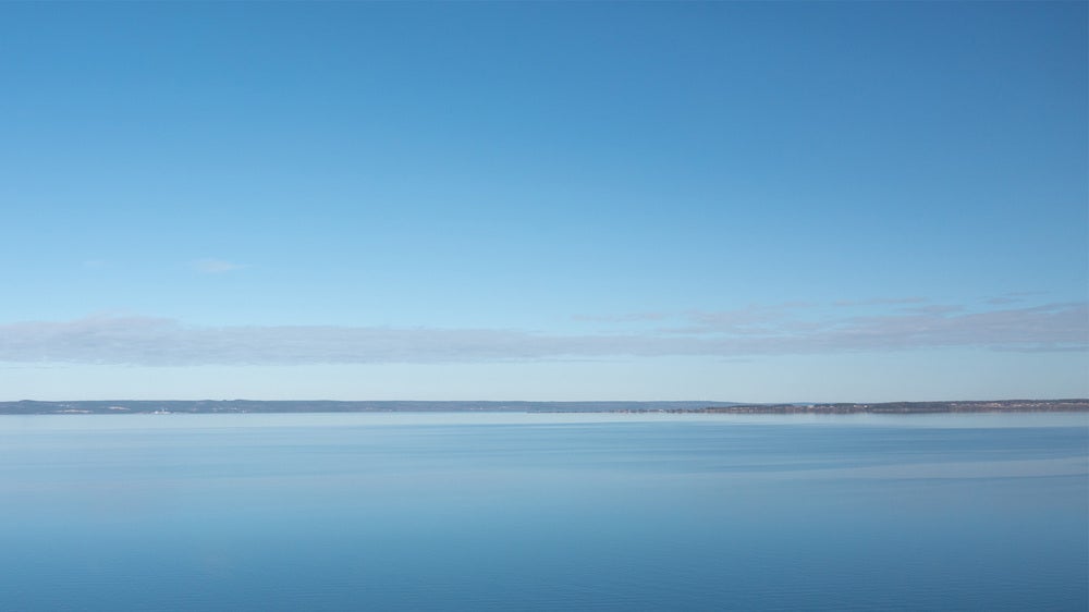 En vidsträckt vy över den lugna sjön Vättern, där himlen och vattnet smälter samman i blå nyanser. Horisonten är tydlig och sträcker sig över hela bilden, vilket skapar en känsla av ro och öppenhet.