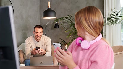 En pappa i ljus tröja sitter i soffan med sin mobil i handen och en laptop framför sig, medan han tittar på sin dotter. Dottern, klädd i en rosa tröja och hörlurar, håller en mobiltelefon och har en skärm framför sig. Båda är uppkopplade och surfar via fibernät i vardagsmiljö.