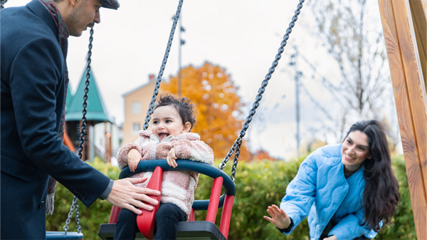 Ett barn gungar glatt medan en vuxen puttar på och en annan vinkar, omgivna av färgglada höstträd i en lekpark.