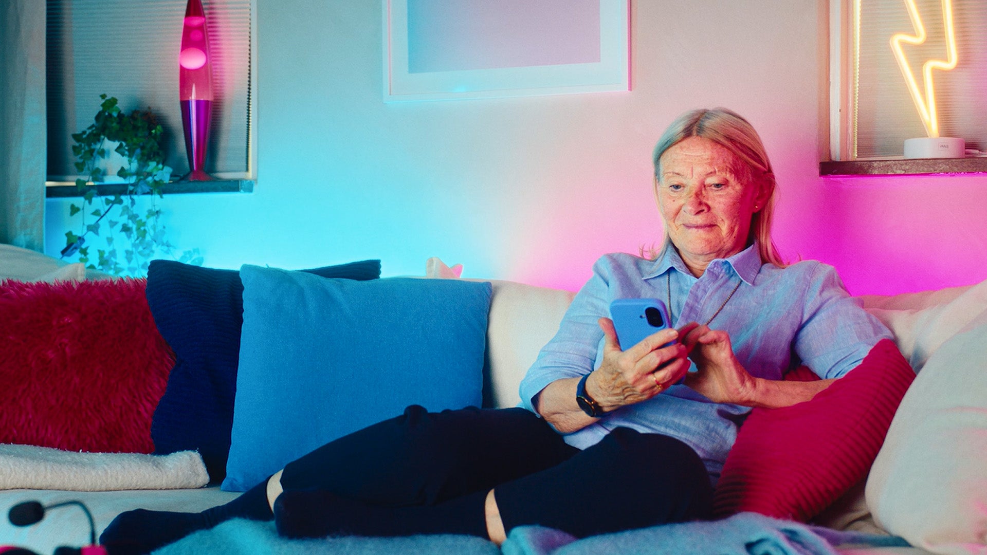 Äldre kvinna med blå skjorta sitter i soffan och trycker på mobilen för att klicka hem ett elhandelsavtal.