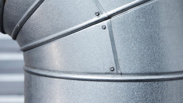 En närbild på ett böjt metallrör i ett industriellt system. Röret är sammanfogat med skruvar och har en slät, galvaniserad yta, vilket ger intryck av en robust installation i en teknisk anläggning.