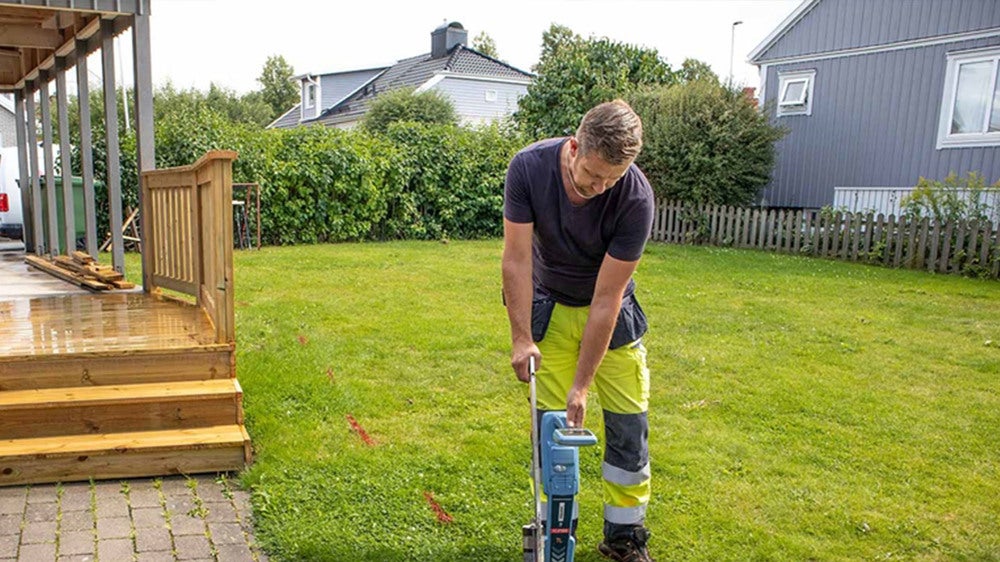 En tekniker iförd arbetskläder markerar ledningsvisning med röda streck på en gräsmatta vid ett bostadshus med trädgård och altan.