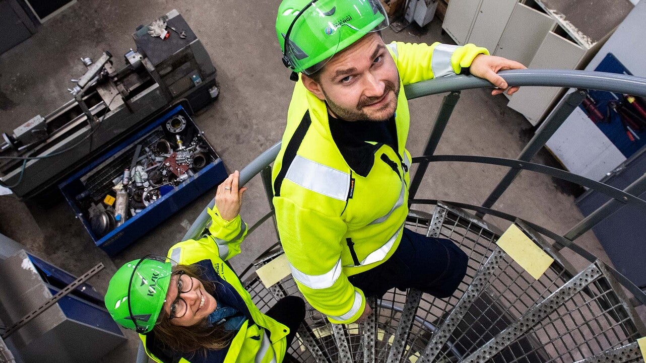 Två tekniker från Jönköping Energi, iklädda gröna hjälmar och gula skyddsjackor, går uppför en spiraltrappa i en industriell miljö. De tittar uppåt och ler, med teknisk utrustning synlig nedanför dem.