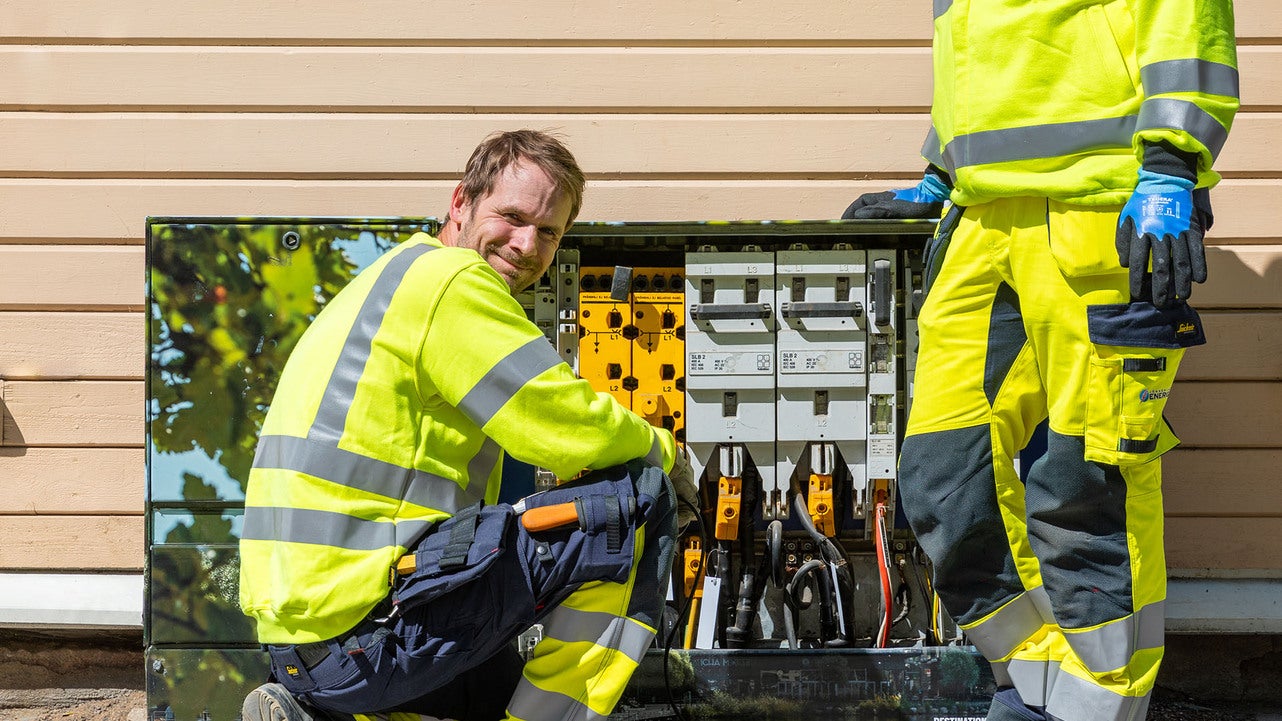 Två tekniker i varselkläder och skyddshandskar arbetar vid ett öppet elskåp utomhus, med en av dem sittande på huk och leende mot kameran.