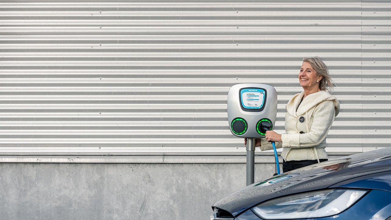En kvinna i ljus jacka laddar sin elbil vid en laddstation utomhus. Hon håller i laddkabeln och ser leende ut medan hon står bredvid bilen. Bakgrunden består av en vägg med metallpaneler.