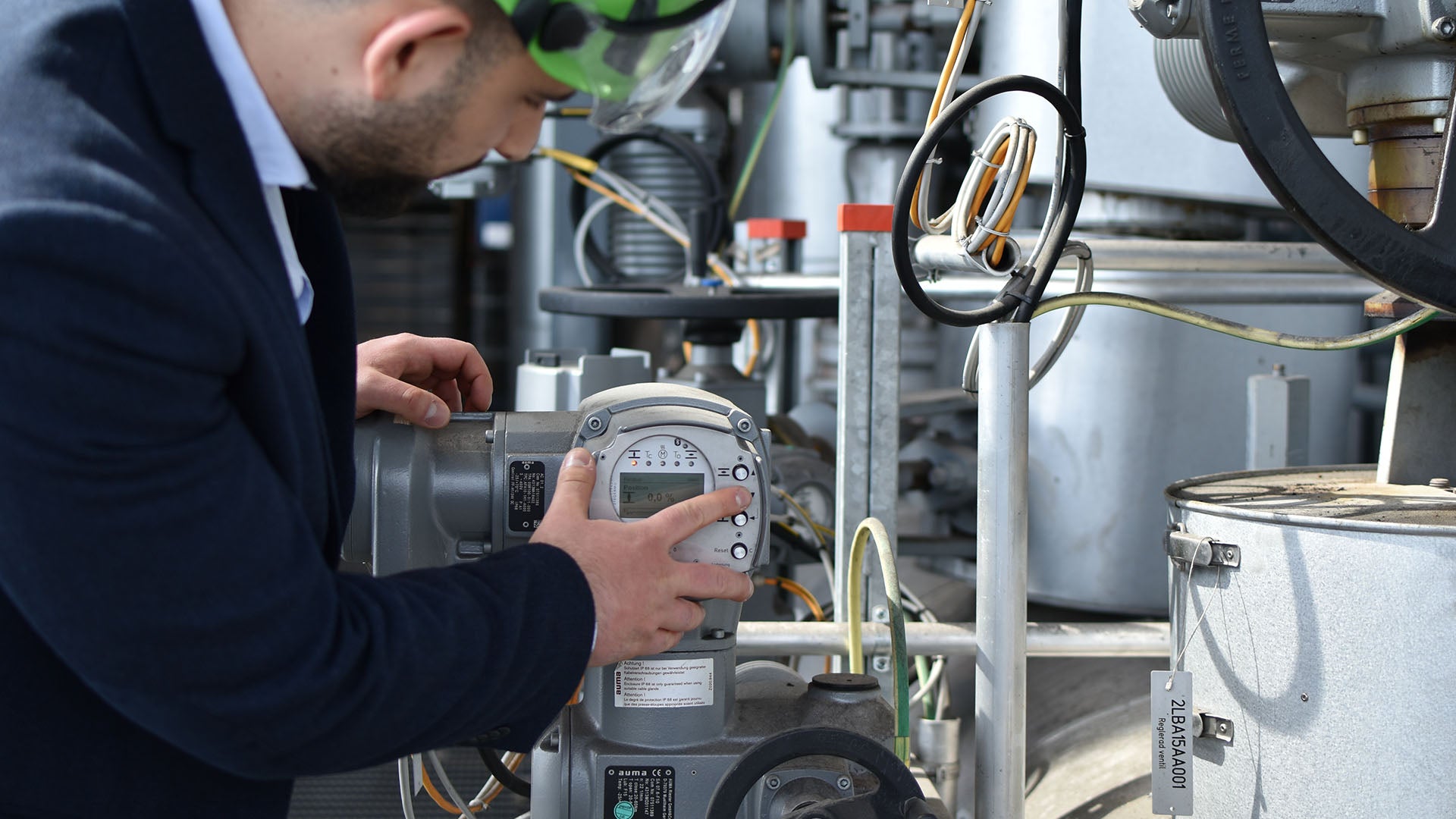 En man i mörk kavaj och skyddshjälm justerar en industriell mätutrustning omgiven av rör och kablar i en teknisk miljö.