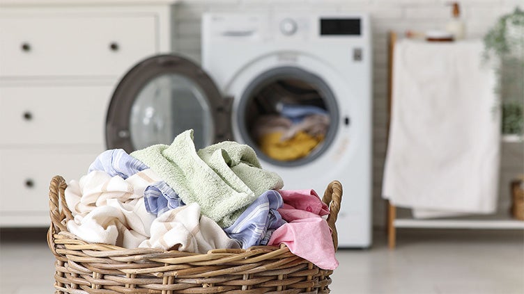 Fylld tvättkorg i förgrunden framför en öppen tvättmaskin i ett ljust tvättrum, med handdukar i bakgrunden.