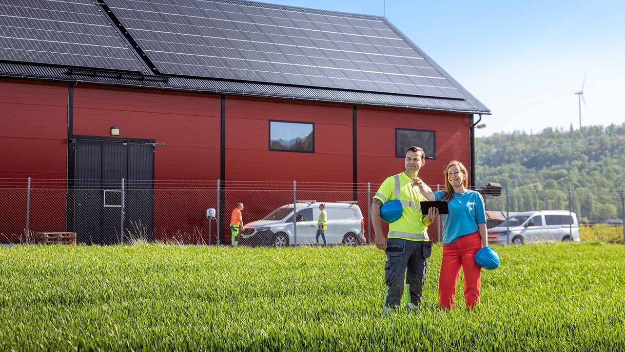 Två personer står framför en röd byggnad med solceller på taket, omgivna av grönska. De håller blå hjälmar och en av dem har en surfplatta. I bakgrunden syns bilar och en vindkraftverk på en kulle.