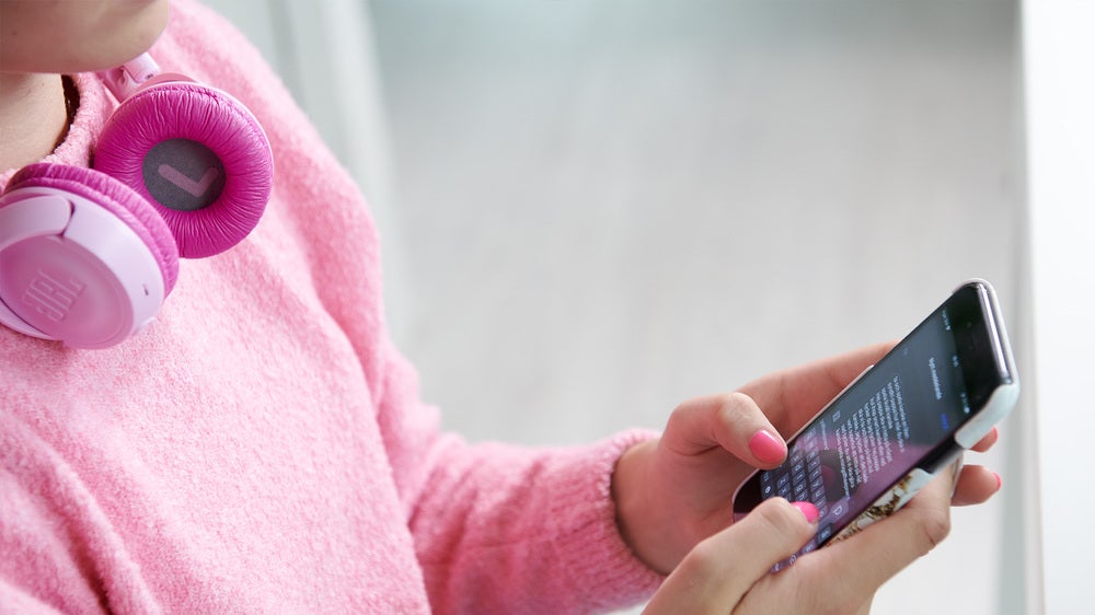 En person i rosa tröja och rosa hörlurar runt halsen använder en smartphone. Personen håller mobilen i båda händerna och tittar på skärmen.