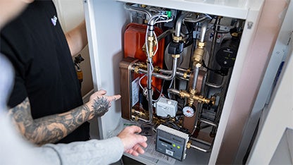 Två personer inspekterar ett öppet värmesystem, där kopparrör, ventiler och en tryckmätare är synliga. Den ena personen har en tatuering på handen och pekar mot systemets delar, medan den andra gestikulerar vid sidan av.