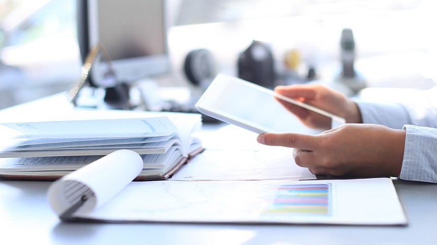 Ett skrivbord med skrivblock, dator och papper. Två händer lutar mot bordet och håller i en iPad.