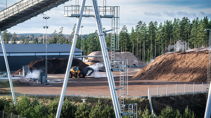 Lastmaskin förflyttar biobränsle vid Jönköping Energis anläggning, omgiven av stora högar med flis och infrastruktur för energiproduktion nära skogsområde.