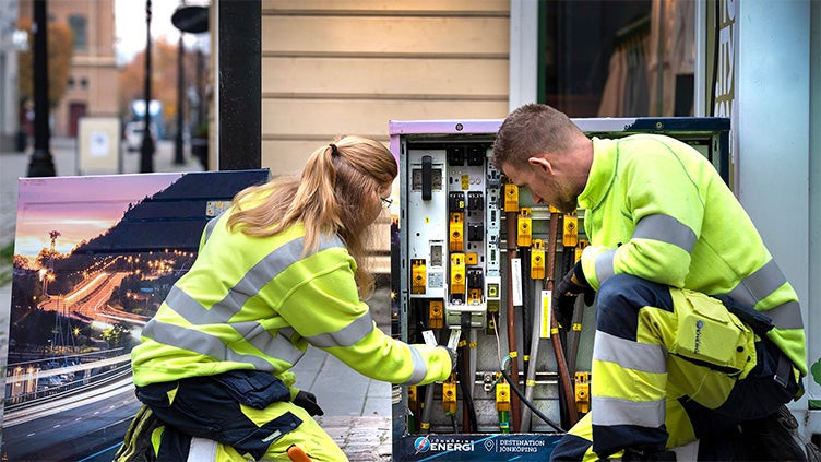 Två tekniker i varselkläder från Jönköping Energi arbetar tillsammans vid ett öppet elskåp på en trottoar i stadsmiljö, med ett reklamskylt i bakgrunden.
