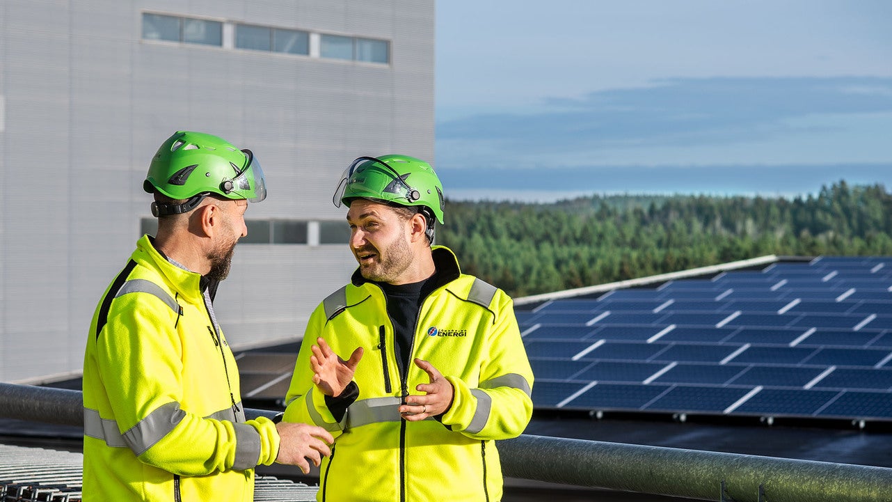 Två tekniker från Jönköping Energi, klädda i gula varselkläder och gröna hjälmar, står framför en solcellsanläggning och har en engagerad diskussion. I bakgrunden syns en industribyggnad och en skogsklädd horisont.