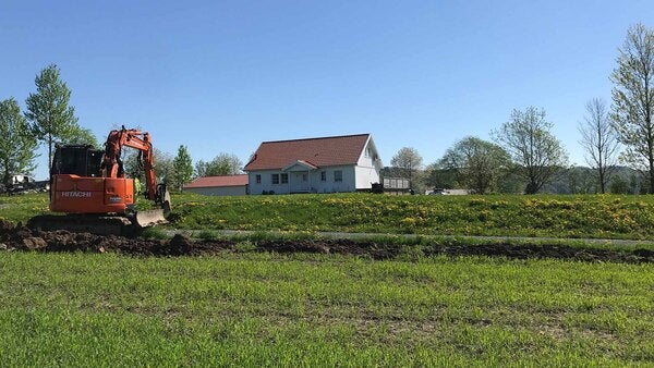 En grävmaskin arbetar med att gräva ned fiberkabel på en grön åker ute på landsbygden. I bakgrunden syns ett vitt hus med rött tak, omgiven av grönska och träd.