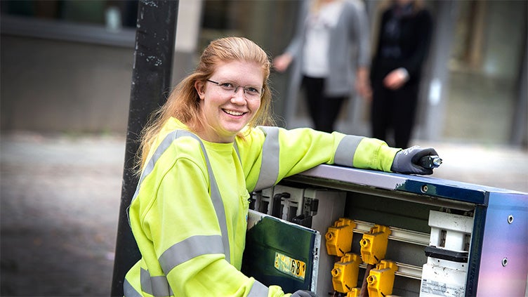 Leende tekniker i varselkläder arbetar vid ett öppet kabelskåp i stadsmiljö, med elkomponenter synliga och förbipasserande i bakgrunden.