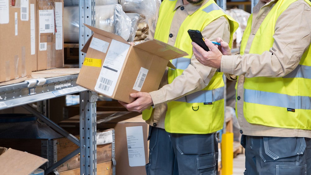 Två lagerarbetare i gula reflexvästar hanterar ett paket framför hyllor fyllda med kartonger. En av dem håller en mobil enhet för att skanna eller kontrollera information om paketet.