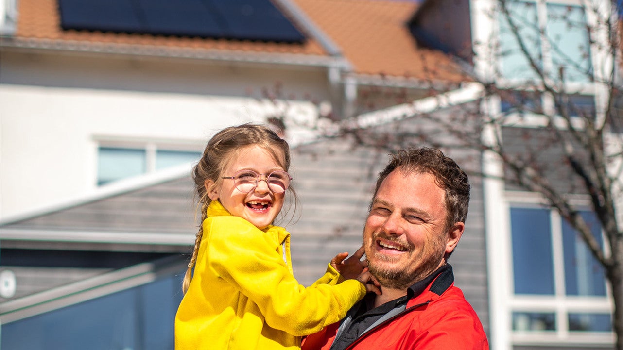 En leende man klädd i röd jacka håller upp sin dotter, som är klädd i gult, framför ett modernt hus med solpaneler på taket. De ser glada ut och solen skiner.