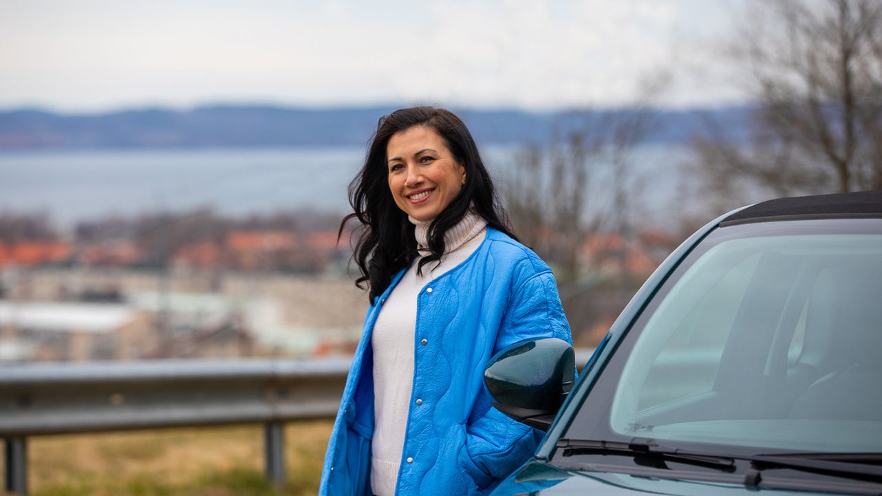 En leende kvinna i blå jacka står vid en elbil med utsikt över Jönköping och Vättern i bakgrunden. Landskapet bakom henne visar både staden och sjön på en mulen dag.