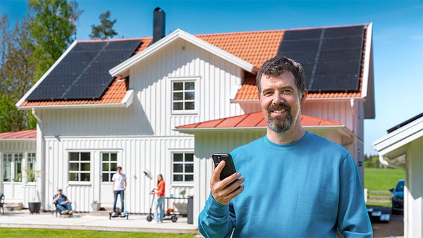 en man i blå tröja håller i en mobil och står framför en stor vit villa med solceller på taket.