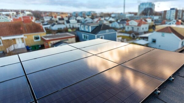 Solpaneler monterade på tak i ett tätbebyggt bostadsområde med flera hus i bakgrunden, fotograferat i dagsljus.