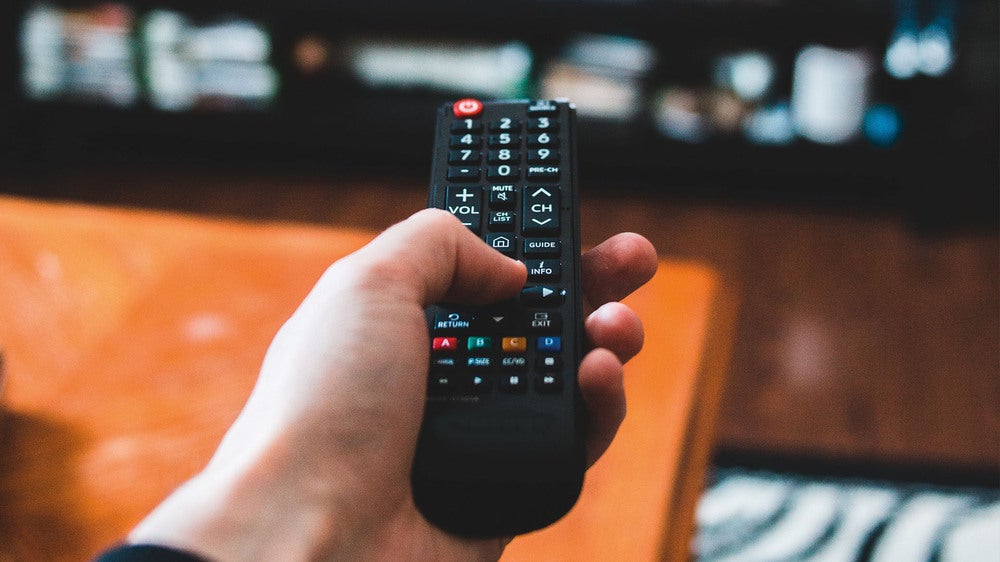 En hand håller i en fjärrkontroll riktad mot en TV eller medieenhet i ett vardagsrum. I bakgrunden syns en suddig TV-bänk och några elektroniska enheter.