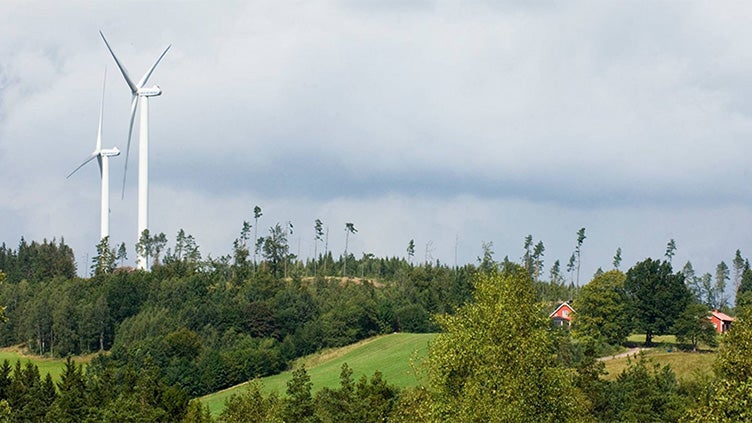 Två vindkraftverk reser sig över ett kuperat skogslandskap med öppna fält och röda hus, under en mulen himmel.