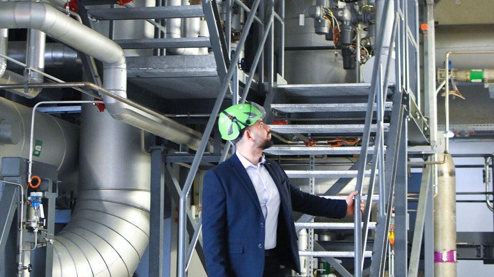 En man i mörk kavaj och skyddshjälm står vid en metalltrappa och inspekterar en industriell anläggning med stora rör och teknisk utrustning.